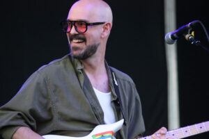 Male guitarist smiling while playing a white electric guitar on stage, wearing a green shirt and glasses.