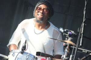Smiling drummer wearing a black knit beanie and orange glasses, in a white mesh shirt, playing a drum set on stage.