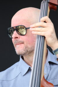 A bald man wearing sunglasses plays a double bass; reflection of a performance visible in the glasses.