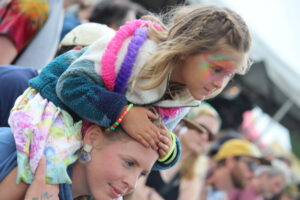 Girl with rainbow face paint riding on another child’s shoulders at a crowded event or parade mountain of people in the background.