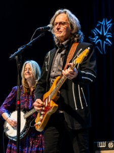 Two musicians perform on stage: a man with a sunburst electric guitar at the foreground and a woman playing a banjo in the background.