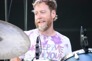 Bearded man smiling while playing drums on stage, cymbals visible in the foreground and a purple tie-dye shirt on his torso.