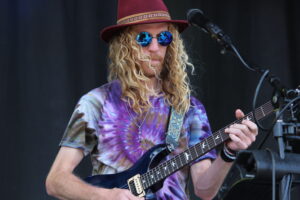 Guitarist with long blond hair, red hat and blue mirrored sunglasses, playing a black electric guitar on stage.