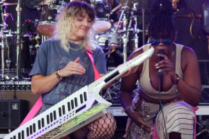 Two female musicians performing on a stage: one plays a white keytar with a pink strap, the other sings into a microphone as drums loom in the background.