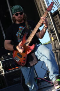 Bass guitarist on an outdoor stage, wearing a green bandana and sunglasses, playing a red sunburst bass.