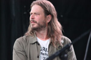 Male musician with long hair and beard on stage, wearing a greenish button-up shirt over a white graphic T-shirt, against a dark background.