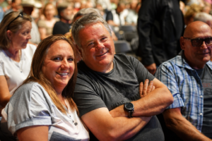 Smiling couple seated at an event, the man in a gray T-shirt with a wristwatch and the woman in a light-colored top, crowd in the background