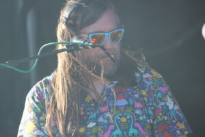 Person performing on stage wearing a bright floral shirt and blue sunglasses, singing into a microphone.