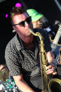 Saxophonist wearing sunglasses and a patterned shirt, playing a golden sax on stage with purple lights in the background.