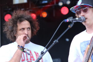Two male performers on stage during a concert: one with curly hair smoking, the other wearing a colorful hat and singing into a microphone.