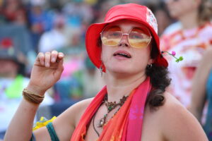 Close-up of a festival-goer wearing a red hat and orange-tinted sunglasses with reflective lenses and layered jewelry.