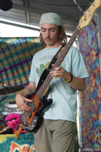 Male bass guitarist wearing a light blue shirt and a light patterned bandana, playing a sunburst bass on a colorful tie-dye stage backdrop.