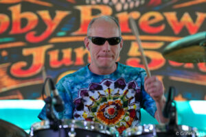 Drummer wearing sunglasses and a tie-dye shirt playing drums on a colorful stage backdrop behind him.