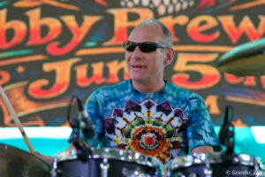Drummer wearing dark sunglasses and a colorful tie-dye shirt plays drums on a stage with a bright, abstract backdrop behind him.