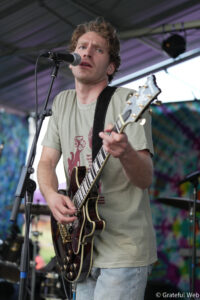 Male guitarist singing into a microphone while playing a sunburst electric guitar on an outdoor stage.