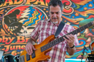 Musician playing a wooden bass guitar on a colorful mural backdrop; focus on the guitarist's plaid shirt and instrument straps