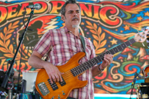Male bassist on stage playing an orange bass guitar, with a colorful swirling mural behind him.