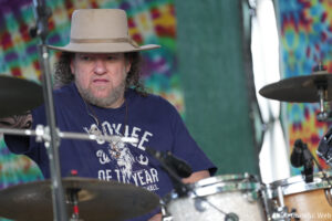 Drummer wearing a beige hat playing drums on a colorful backdrop, close-up of his face and kit in focus