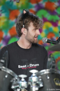 Man wearing a cat-ear headband singing into a microphone at a drum set, colorful background behind him