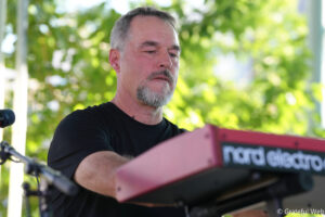 Man with gray beard playing a red keyboard outdoors, microphone nearby, green foliage in the background.