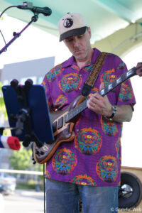 Male guitarist wearing a beige cap and a purple patterned shirt plays a brown electric guitar on an outdoor stage, with a microphone nearby.