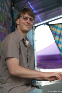 Man with glasses smiling as he plays a keyboard, with a colorful mural and purple lights in the background