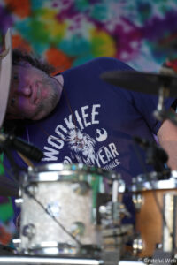 Bearded man in a beige hat playing drums, seen from the side against a bright tie-dye backdrop.
