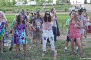Group of people dancing at an outdoor festival in a field, wearing colorful tie-dye clothing and summer outfits.