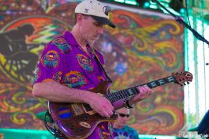 Man in a purple paisley shirt and white cap playing an electric guitar on a vibrant psychedelic stage backdrop.