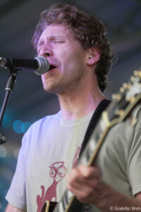 Musician singing into a microphone while playing an electric guitar on stage, wearing a light T-shirt.