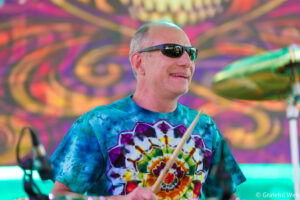 Drummer wearing sunglasses and a tie-dye shirt plays drums against a bright, psychedelic backdrop.