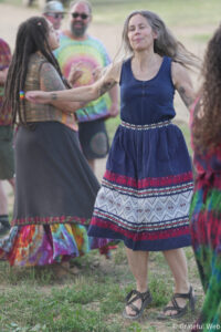 Group of people dancing outdoors, woman in a blue patterned dress in the foreground, others in colorful clothing behind her.