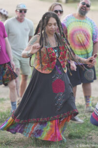 Woman with long dreadlocks dancing in a colorful floral-embroidered dress at an outdoor festival, surrounded by friends in casual, tie-dyed clothing.