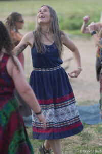 Woman in a blue patterned dress dancing outdoors with friends, smiling up at the sky.