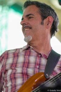 Side profile of a smiling man with a gray goatee wearing a plaid shirt, playing an electric guitar with a strap visible