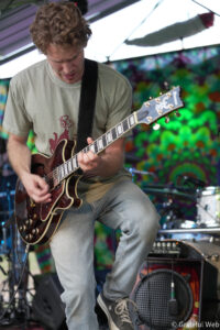 Guitarist playing a dark red electric guitar on stage with a colorful mural backdrop behind him.