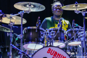 Drummer on stage behind a large drum kit with cymbals, wearing a green and yellow shirt, under blue stage lights.