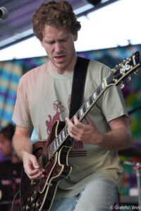 Male guitarist performing live on stage, concentrating while playing a dark red electric guitar with a strap over a beige T-shirt