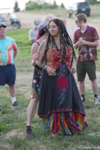 Woman with long dreadlocks in a floral dress stands in a grassy field at an outdoor gathering, smiling with friends behind her.