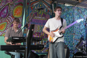 Two musicians perform on a colorful tie-dye stage backdrop; keyboardist on the left and guitarist on the right playing a sunburst Fender-style guitar, with drums nearby.