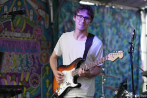 Young man with glasses playing an electric guitar on a colorful graffiti backdrop stage.