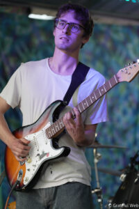 Young man with glasses playing a sunburst Fender Stratocaster on stage under purple lighting.