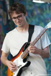 Young man with glasses playing a sunburst electric guitar, looking down at the fretboard.