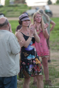 Woman wearing a lavender cap and a colorful mushroom-pattern dress, clapping at an outdoor gathering.