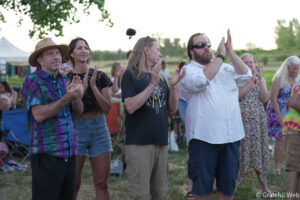 Group of adults outdoors, standing and clapping at an event with tents and trees in the background.