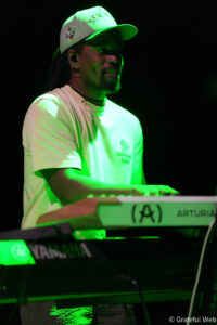 Keyboardist performing on stage under green lights, wearing a white cap and T‑shirt.