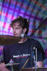 Drummer wearing glittery cat-ear headband playing drums on a colorful blurred backdrop.