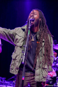 Male singer with long dreadlocks performing on stage into a microphone under purple lights