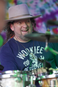 Smiling drummer wearing a beige wide-brim hat while playing a drum kit, colorful backdrop behind him.