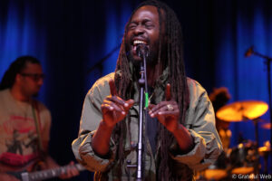 Male singer with long dreadlocks, wearing a green jacket, singing into a microphone on a blue-lit stage, hands raised in front.
